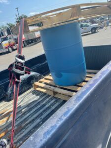 Wooden crate flooring placed on the top and bottom of a blue shipping drum strapped to the back of a pickup truck